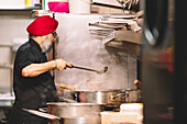 A chef in a red turban stirring a large pot in a steamy, commercial kitchen