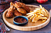 Crispy fried chicken hips alongside golden french fries and a dipping bowl of barbecue sauce, served on a round wooden platter.