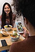 From From above of unrecognizable woman taking photo of poke dish on smartphone while spending weekend in restaurant with female friend