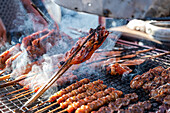 Juicy chicken skewers sizzle over charcoal, with smoke rising amid the busy food stall in Bangkok, Thailand, showcasing traditional street food.