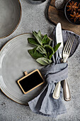 This image captures a stylish summer table setting featuring a ceramic plate, silver cutlery wrapped in a gray linen napkin, and fresh mint leaves, all arranged on a textured surface