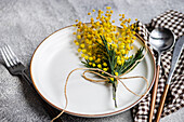 A rustic table setting featuring a white plate with a rim, accented with a sprig of mimosa flowers tied with twine, alongside silverware and checkered napkin