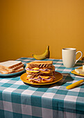 Ein hausgemachtes Frühstück auf einer blau karierten Tischdecke mit Sandwich, Banane, Kaffee und Butter vor gelbem Hintergrund