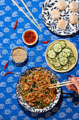 Top view of an assortment of Chinese dishes including shrimp noodles, cucumber salad, dumplings, and sauces. A hand is seen picking up noodles with chopsticks, enhancing the lively dining atmosphere