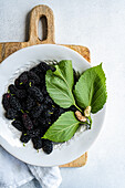 Top view of a white ceramic bowl filled with juicy black mulberries, adorned with vibrant green leaves, rests on a rustic wooden cutting board against a soft grey background. The setup is captured from above, showcasing the rich texture and natural appeal of the fruit, making it an ideal image for food and nutrition themes.