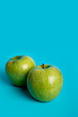 Two vibrant green apples sit against a stark turquoise backdrop, showcasing their glossy texture and natural freshness. This image emphasizes the beauty and simplicity of wholesome food.