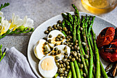 A nutritious meal featuring boiled eggs, barbecued asparagus, capers, and grilled vegetables including tomatoes and chili peppers, artistically arranged on a plate