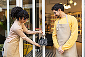 A female business owner unlocks the door of her shop, handing over an "Abierto" sign to a pleased male employee in an apron, signaling the start of the day.