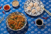 From above, a vibrant display of Chinese food on a blue floral tabletop with shrimp noodles, steamed dumplings in a patterned bowl, and condiments like soy sauce and chili oil