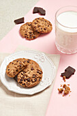 Top view of delicious chocolate chip cookies on a white plate with a glass of milk and scattered chocolate pieces on a pink background