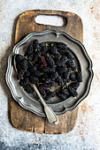 Top view of a rustic setting showcases fresh juicy mulberries in a vintage silver tray, resting on a weathered wooden chopping board, complemented by a textured silver spoon and a dusty backdrop.