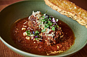 A carefully crafted dish displaying succulent meatballs in a rich tomato sauce, garnished with grated cheese and microgreens, accompanied by a golden bruschetta.