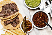 A top view of a traditional Mexican homemade meal featuring grilled steak on corn tortillas, accompanied by guacamole, salsa roja, caramelized onions, and a side of fries.