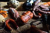 A selection of fresh and cured sausages, including bright red sobrasada, displayed on a worn wooden table, accompanied by rustic cooking tools and spices.