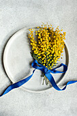 From above of charming mimosa flower sprig bound with a vivid blue ribbon, laid upon a textured table surface, creating a refreshing visual contrast