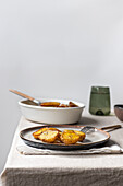 Half-cut roasted potatoes garnished with herbs presented on a ceramic plate alongside a bowl with a wooden spoon and a green cup, all arranged on a linen tablecloth.