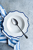 An elegant white plate with scalloped blue edges accompanied by a vintage spoon, set on a concrete background, wrapped in a striped blue tea towel