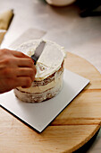 Eine Person glasiert eine mehrschichtige Torte mit cremigem Zuckerguss auf einem Holzbrett mit einem Spachtel, der das Backen zu Hause darstellt