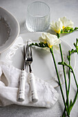 An elegant table with white porcelain dinnerware, polished silverware wrapped in a linen napkin, a textured glass and delicate freesia flowers enhance the refined atmosphere