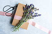 Elegant summer table setting concept highlighted by fresh lavender flowers, neatly presented beside a gift box wrapped in brown paper and a pink ribbon, with vintage scissors on a textured grey surface