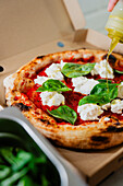 An anonymous person drizzles olive oil on a Margherita pizza topped with mozzarella and basil, freshly baked and served in a cardboard takeout box