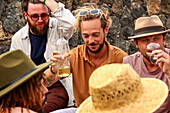 Männer lachen und trinken gemeinsam Wein an einem Tisch im Freien, wobei einer mit Begeisterung Wein in Gläser einschenkt. Alle Männer sind beschäftigt und schauen sich gegenseitig oder die Gläser an.