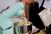 A person baking a cake pours flour into a mixing bowl as a teal-colored modern mixer blends the ingredients together