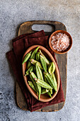 Frische Okra-Bamia-Schoten werden in einer Terrakotta-Schale auf einem hölzernen Schneidebrett vor einem strukturierten grauen Hintergrund präsentiert und verkörpern den rustikalen Charme, der für die mediterrane Küche typisch ist