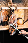 A cropped unrecognizable hand using tongs to pick a rustic sourdough bread loaf from a warm-hued selection displayed on a wooden shelf.