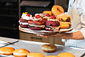 Crop anonymous female baker in uniform standing in bakery with tray of tasty vegan Berliners with whipped cream filling