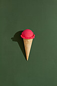 A vibrant Strawberry ice cream scoop sits atop a classic waffle cone against a green background, casting a playful shadow