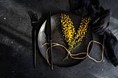 Top view of sophisticated black dinner plate and cutlery set adorned with bright yellow mimosa blossoms, tied with twine, on a dark textured background.