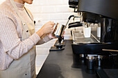 A focused barista using a modern espresso machine to steam milk in a metal pitcher, preparing for a creamy coffee drink.