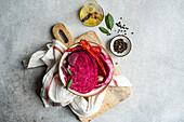 Vibrant beetroot fermented cabbage with spicy red peppers and garlic, served in a bowl on a rustic wooden cutting board, accompanied by olive oil, bay leaves, and whole peppercorns.