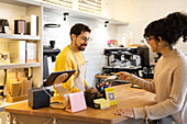 A smiling male barista in a yellow shirt accepts payment from a female customer using a card reader at a modern coffee shop counter.