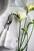 A sophisticated table setting featuring white porcelain plates, silver cutlery with white handles, a linen napkin, and fresh white freesias as centerpieces, perfect for celebratory events