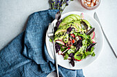 Colorful vegetable salad featuring lettuce, olives, cherry tomatoes, cucumber slices, radishes, red basil, sprinkled with white and black sesame seeds, artfully presented in a white bowl with a blue napkin.