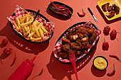 A delectable arrangement of crispy fried chicken and golden fries served in black baskets with red and white checkered liners, surrounded by fresh red chili peppers, mustard, and ketchup, all on a red background