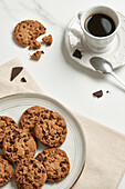 A sophisticated arrangement featuring a cup of black coffee and chocolate chip cookies on a ceramic plate, with dark chocolate shards nearby