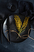 Top view of an artistic table setting featuring modern black tableware and vibrant yellow mimosa flowers, tied with twine for a chic contrast.