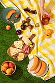 An overhead view of a summer picnic, displaying a colorful array of fresh fruits, cheese, crackers, and sliced meats on a checkered yellow tablecloth, with anonymous hand reaching for a drink.