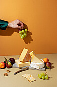 A variety of gourmet cheeses accompanied by fresh fruits, nuts, and crackers on an earth-toned backdrop, highlighted by anonymous person adding grapes to the composition