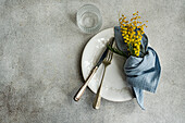 Elegant dining setup featuring a white ornate plate, vintage silverware, a blue linen napkin, and a bright yellow mimosa bouquet on a textured backdrop