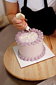 A baker decorates a cake with a pastry bag, adding elegant lettering to the smooth frosted surface