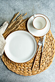 From above view of a stylish table setting featuring a bright white ceramic dinner plate, cup, and sleek silverware atop a woven placemat, accompanied by decorative wheat ears.