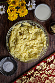 Draufsicht auf einen rustikalen Holztisch mit einer Schüssel mit hausgemachtem Kartoffelpüree, einer kleinen Schale mit Butter, einem Glas Milch und dekorativen Blumen, auf dem ein traditionelles hausgemachtes Mittagessen steht.