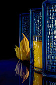 A vibrant glass of mango and lime juice beside a unique Buddha hand fruit, against a textured blue backdrop with a reflective surface