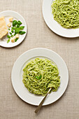 Overhead view of a spaghetti pesto dish on a white plate, accompanied by parmesan and basil leaves.