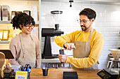 Two cheerful multiethnic baristas in aprons are happily engaging in their duties at a modern, well-lit coffee house, showcasing teamwork and customer service.