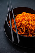 A close-up shot of a spicy Korean carrot salad, served in a black ceramic bowl with chopsticks on a dark background